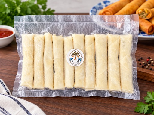 A sealed pack of frozen Asliya Foods spring rolls on a walnut table, with a plate of cooked golden rolls behind it
